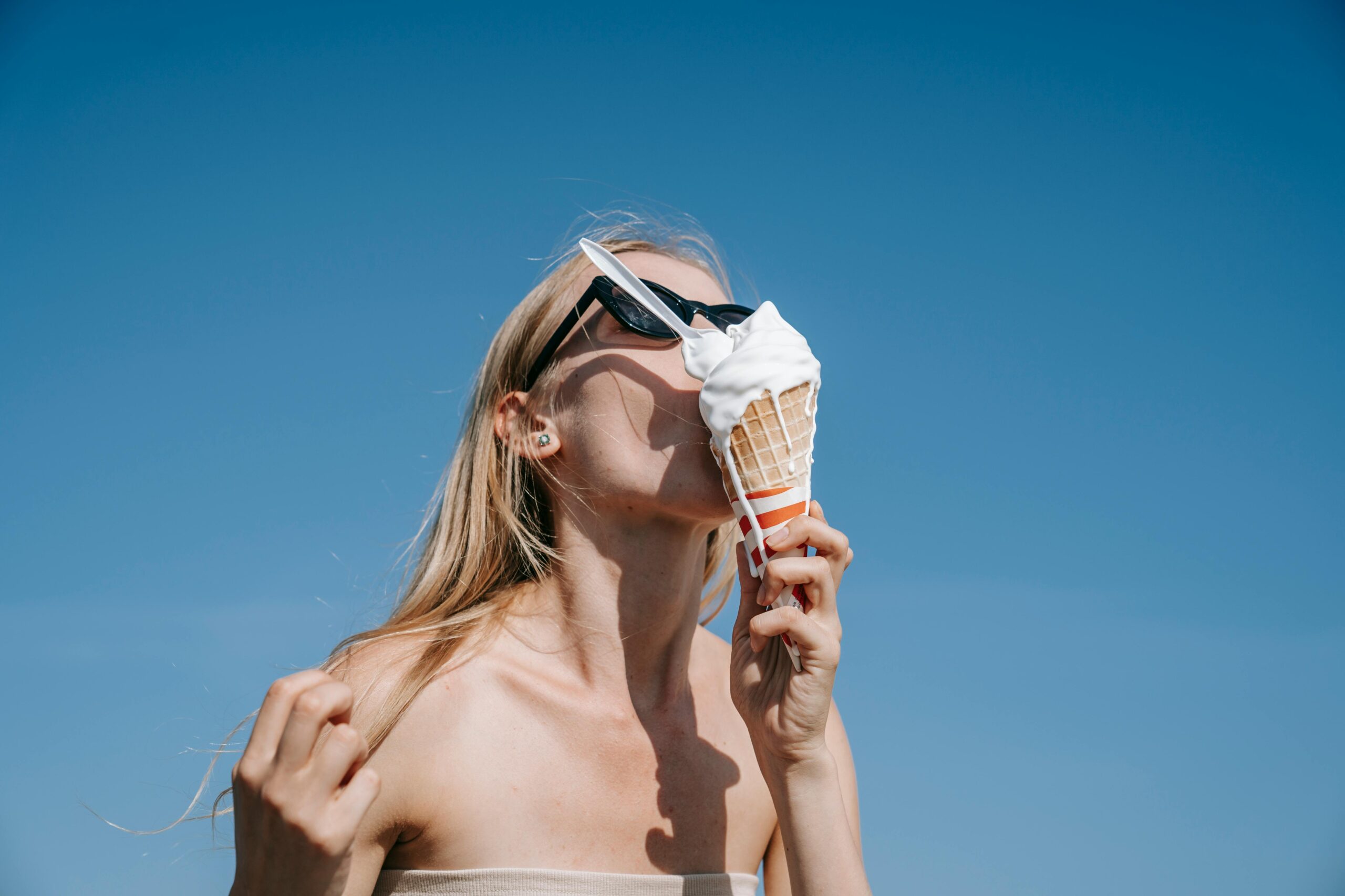 A woman in sunglasses enjoys a melting ice cream cone under a vibrant blue sky.